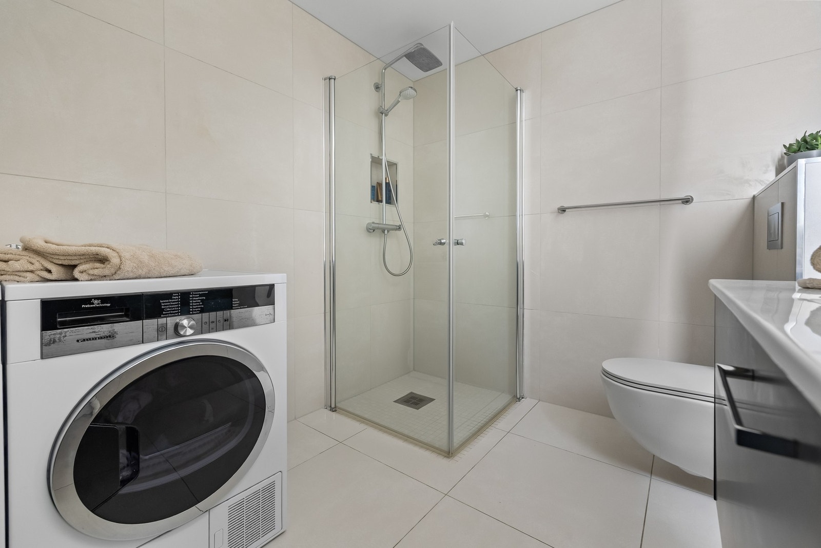 Videre er badet utstyrt med vegghengt WC og en innbydende dusjsone med rainshower, hånddusj og dusjvegger i herdet glass. Det er også tilrettelagt med opplegg for vaskemaskin og tørketrommel. Badet har mekanisk ventilasjon via ventil i himling, og med til Galleribilde