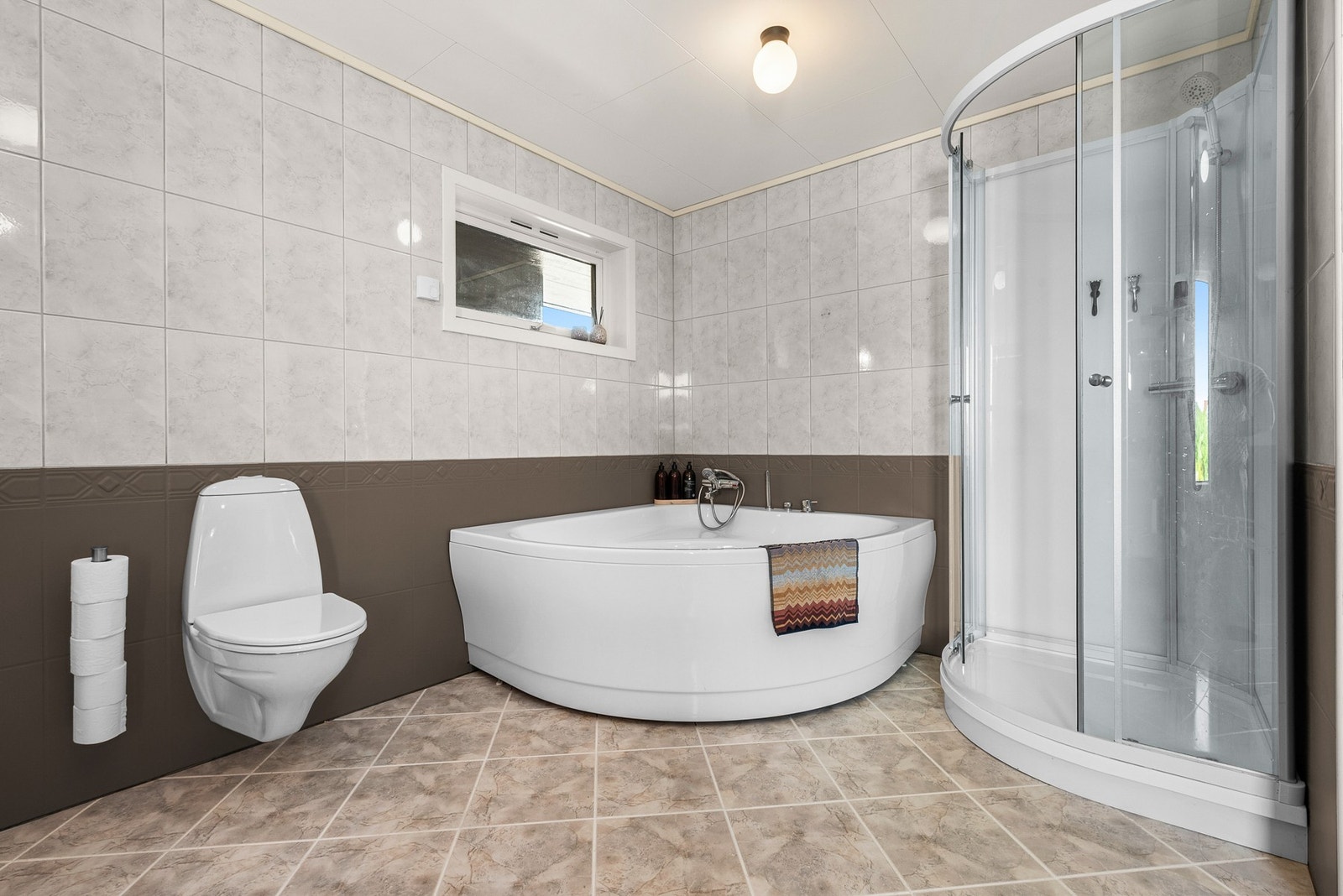 Badet har flis på gulv og vegger, badekar/dusjkabinett, veggmontert toalett og servant i innredning. Galleribilde