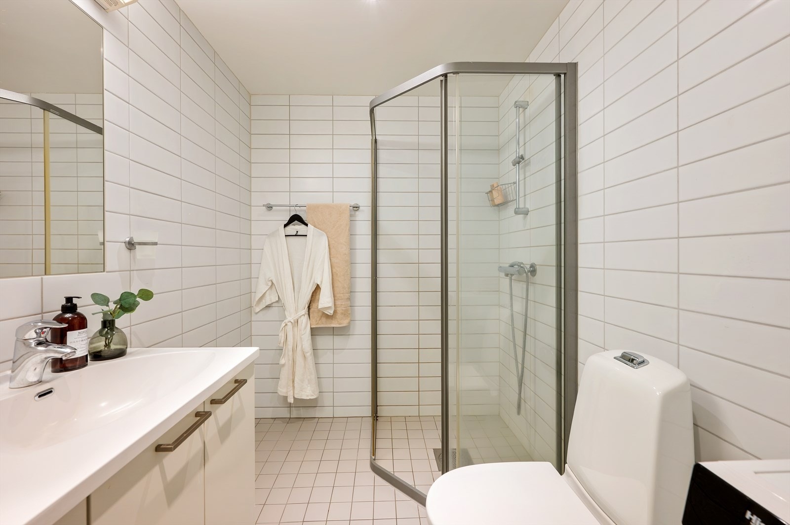 Badet fremstår som lyst og funksjonelt med et tidløst uttrykk. Rommet er helfliset med fliser på vegger og gulv, og har varmekabler i gulv som gir god komfort. Innredningen består av servant med underskap og speil, samt toalett og dusjhjørne Galleribilde