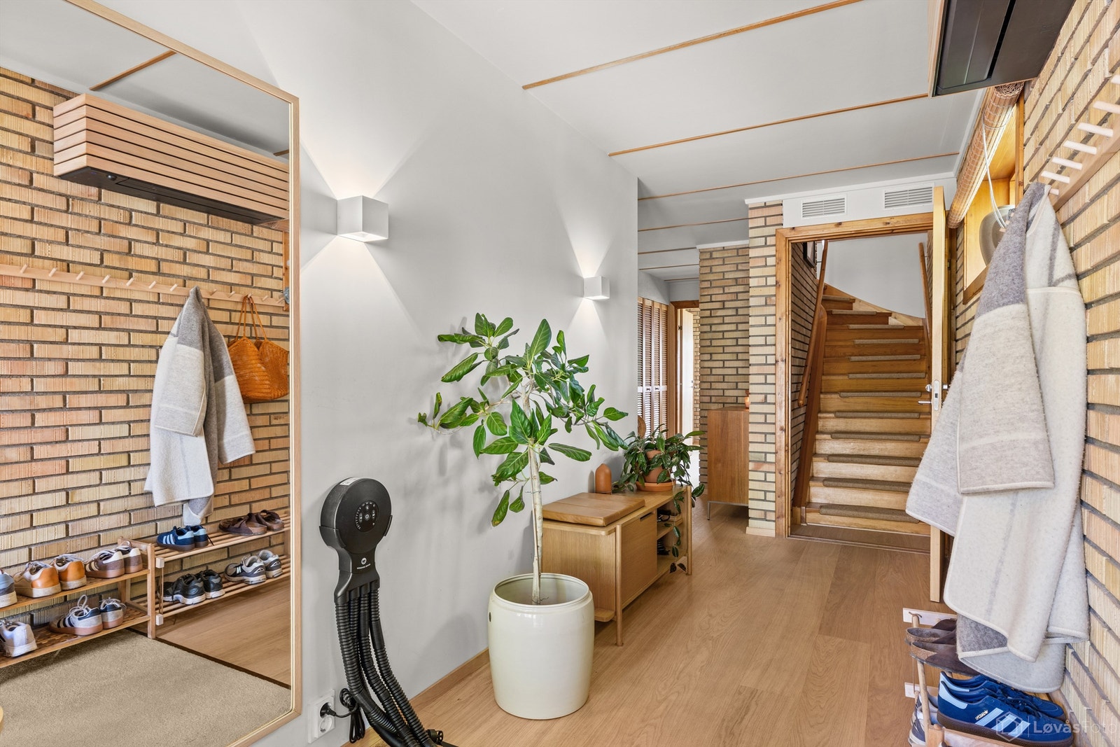 Romslig hall med eksponert teglmur på den ene siden. Hallen fungerer som knutepunkt for hele boligen - soverom, bad og wc ligger i samme plan, mens de sosiale rommene venter en etasje opp. Galleribilde
