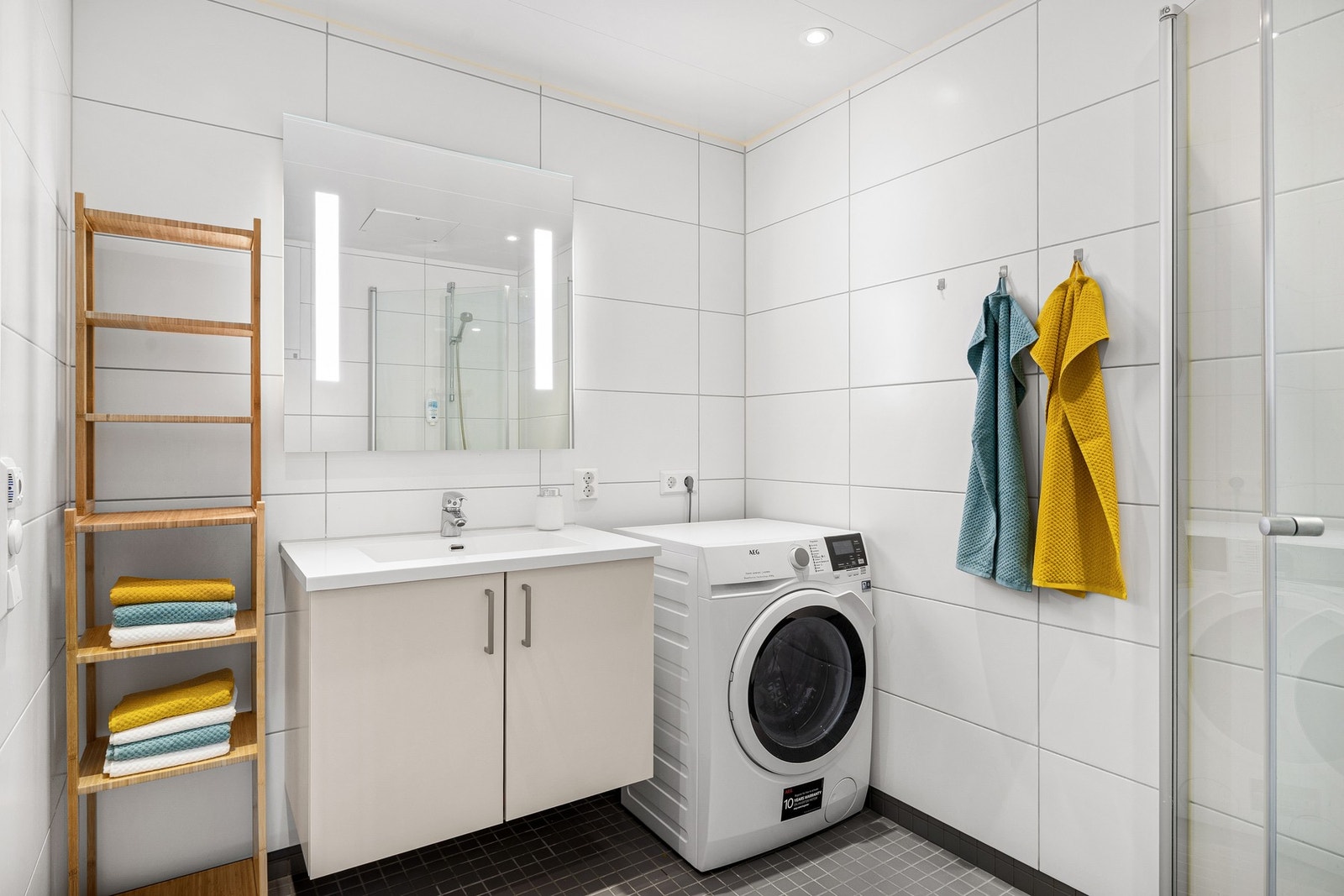 Det lyse baderommet inneholder innfellbare og plassbesparende dusjdører i glass, dusjbatteri og garnityr montert på vegg, veggmontert WC og kran og avløp for vaskemaskin. Badet varmes opp ved bruk av elektriske varmekabler. Galleribilde