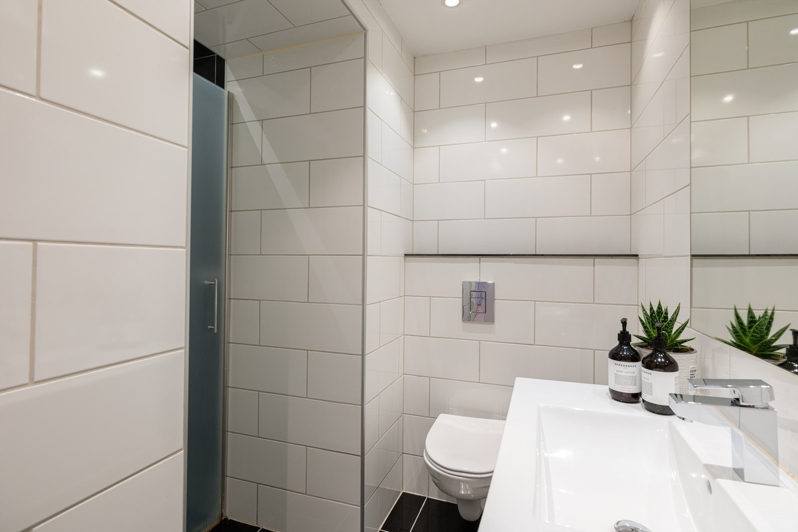 Badet har innfelte downlights i taket og er innredet med vegghengt toalett, servant nedfelt i moderne baderomsinnredning samt dusjhjørne med svingbare glassdører. Galleribilde