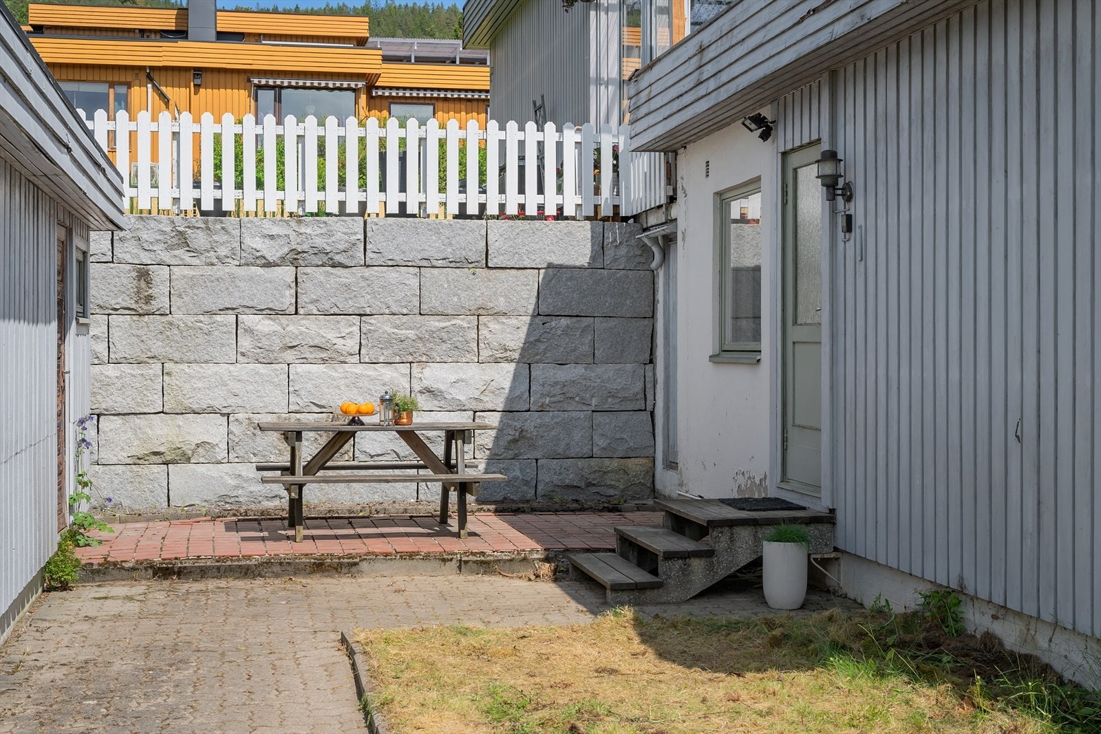 En solid steinmur skaper en fin avgrensning og en lun uteplass på eiendommen. Boligen disponerer en ekstern bod på 3 m² (døra helt til høyre i bildet. Boden ligger under terrassen til naboen over) Galleribilde