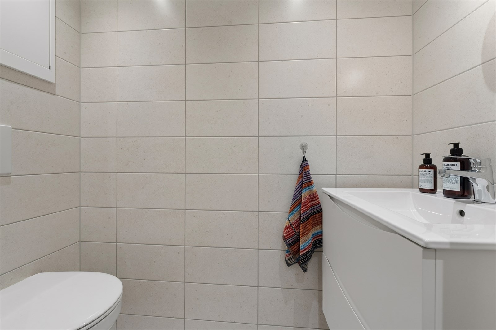 Lyst og moderne toalettrom med flislagte overflater, vegghengt toalett og servant med oppbevaring. Galleribilde