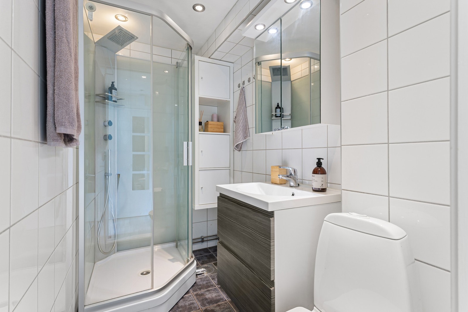 Baderommet med fliser på vegger og gulv inneholder gulvmontert WC, dusjkabinett og servant på underskap. Badet varmes opp ved bruk av elektriske varmekabler. Galleribilde