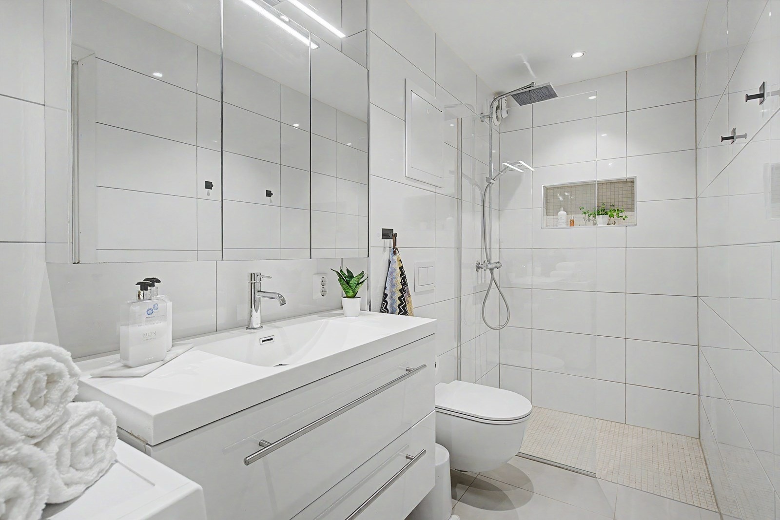Stort og innbydende bad, i tidsløse fargepaletter. Gjennomførte detaljer med fliser på vegger og på gulv, dusjnisje i vegg til oppbevaring av såper. Stor og romslig dusj, med waterfall Galleribilde