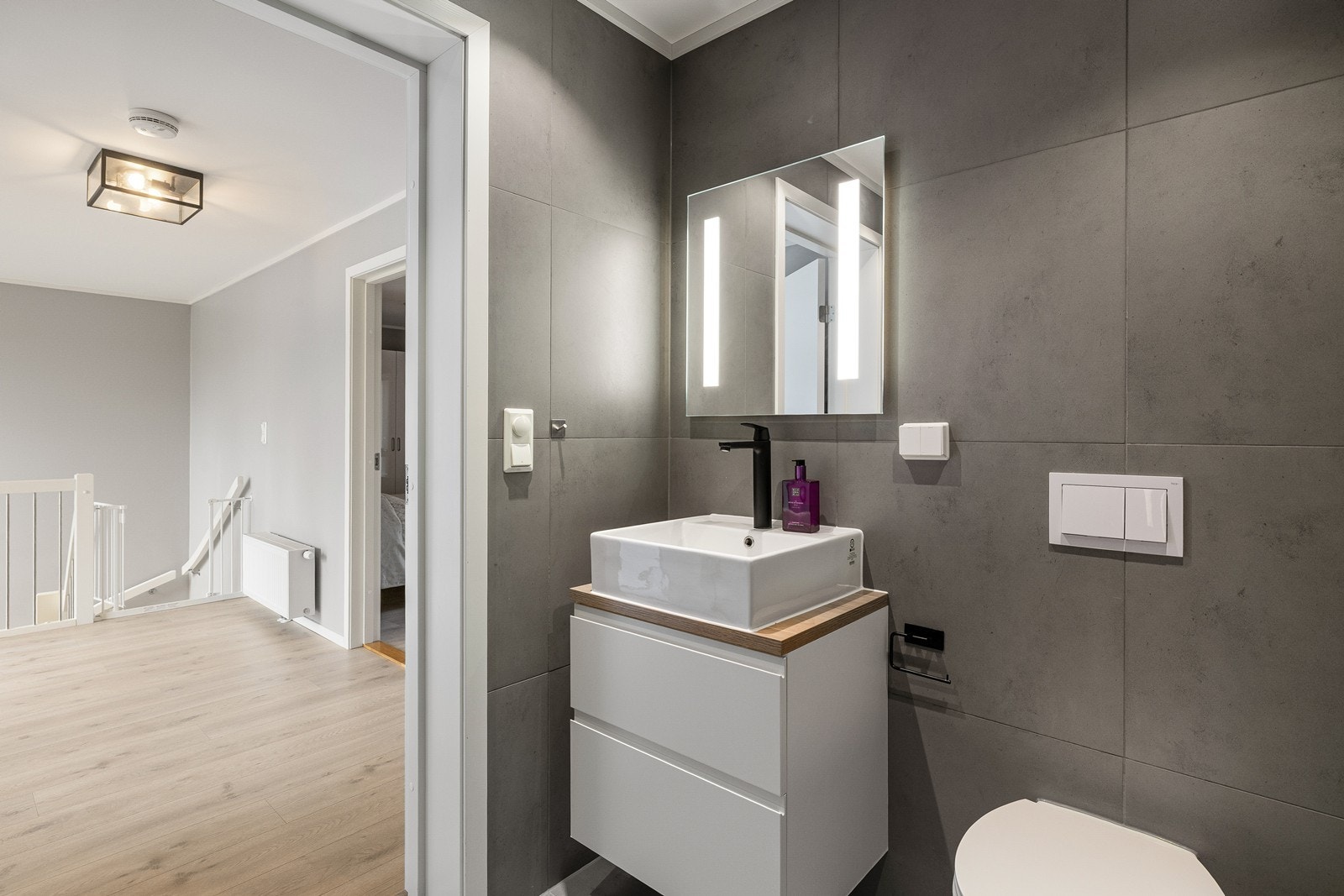 Innredningen består av et minimalistisk, hvitt servantskap med glatte fronter og en delikat toppmontert servant. Over vasken er det montert et stort speil med integrert og behagelig LED-belysning på sidene. Galleribilde