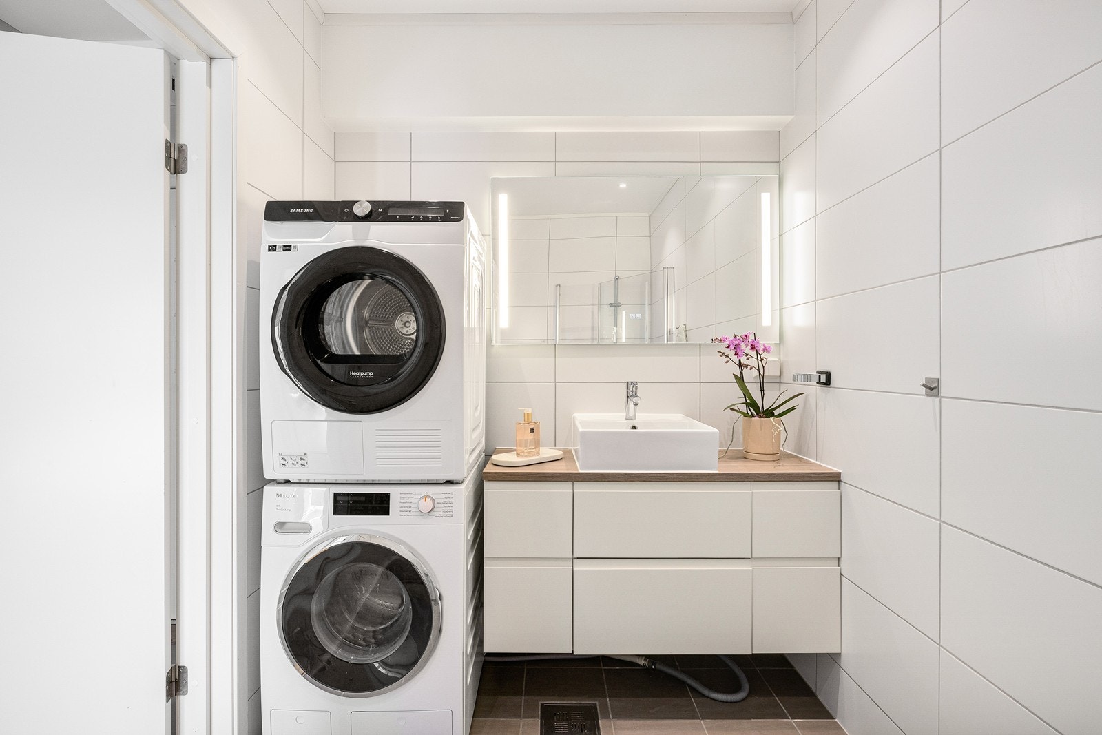 Badet i første etasje fungerer også som et praktisk vaskerom. Dette er et stilrent bad utstyrt med servantskap, speil med lys, vegghengt toalett, dusjhjørne og opplegg for vaskemaskin. Galleribilde