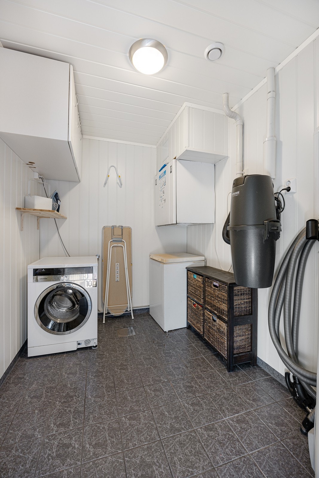 Vaskerommet ligger vegg-i-vegg med badet, og har hvitmalte vegger og grå fliser på gulv med gulvvame. Galleribilde