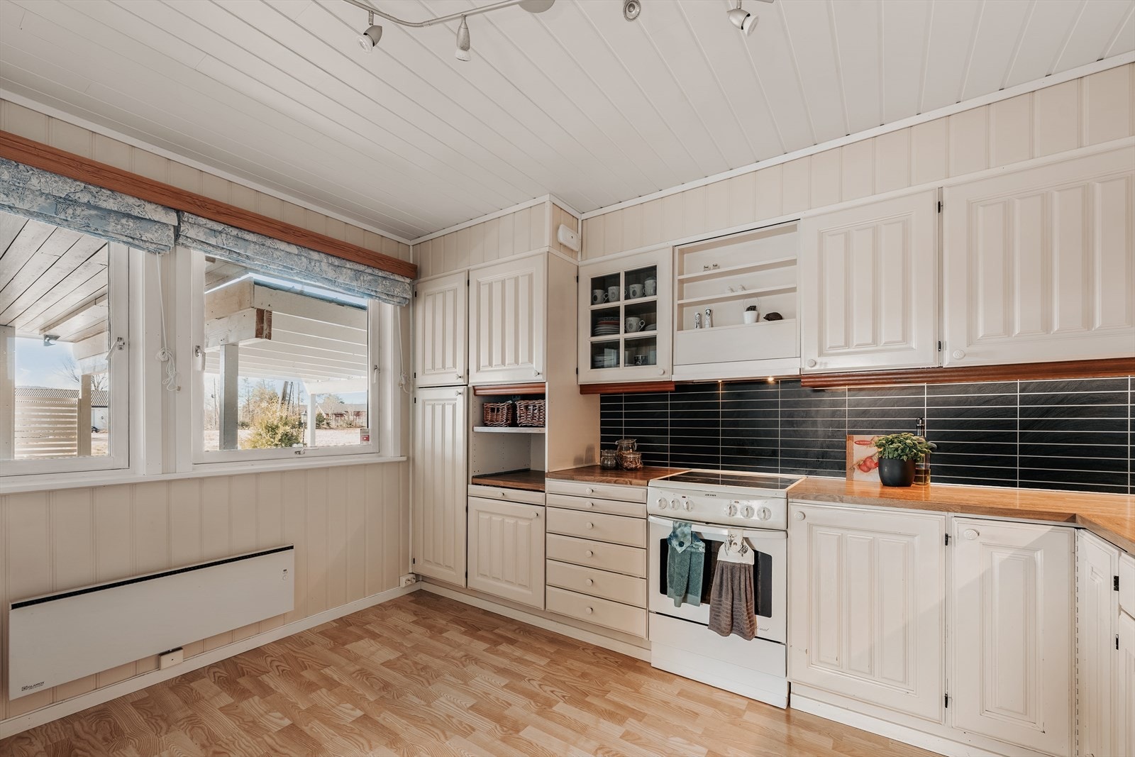 Separat kjøkken med en eldre kjøkkeninnredning med profilerte trefronter, benkeplate i treverk med nedfelt kum i rustfritt stål og plater på vegg over benkeplate. Galleribilde