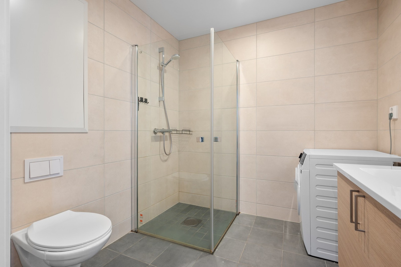 Badet er innredet med innfellbare dusjdører i glass, veggmontert WC, opplegg for vaskemaskin, servant på underskap, og speil. Galleribilde