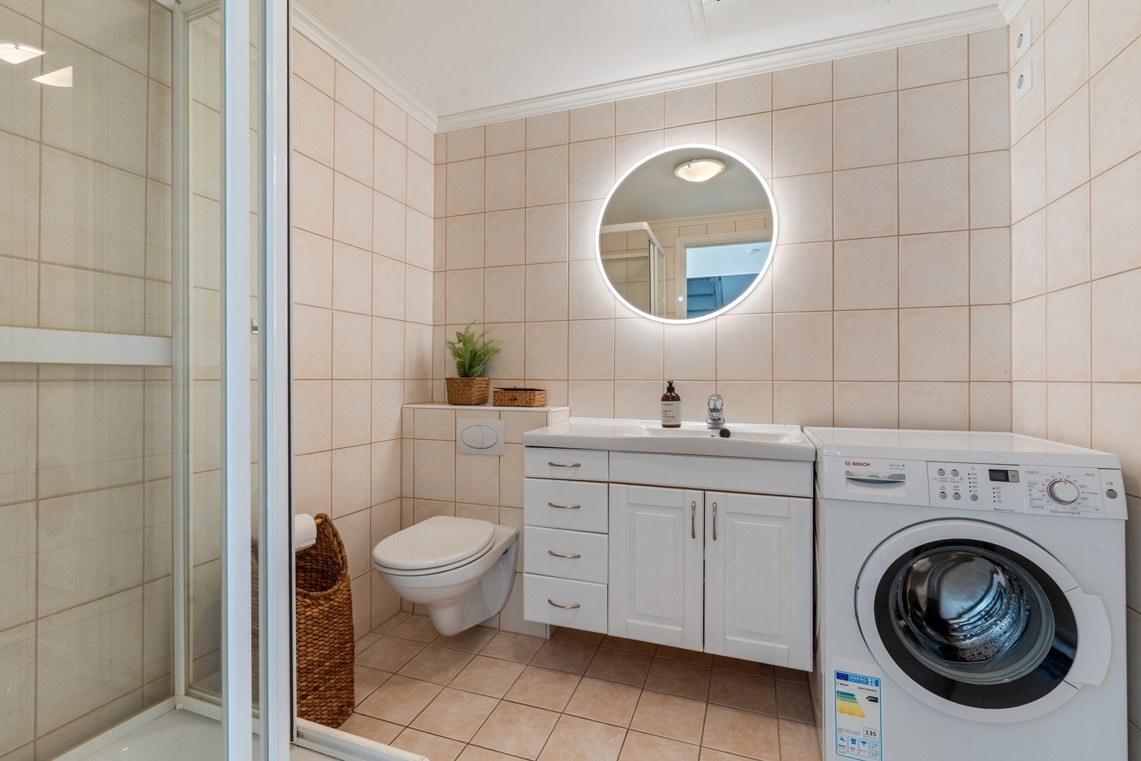Badet er utstyrt med servant i møblement, speil med belysning, vegghengt toalett, dusjkabinett og opplegg til tørk/vask Galleribilde