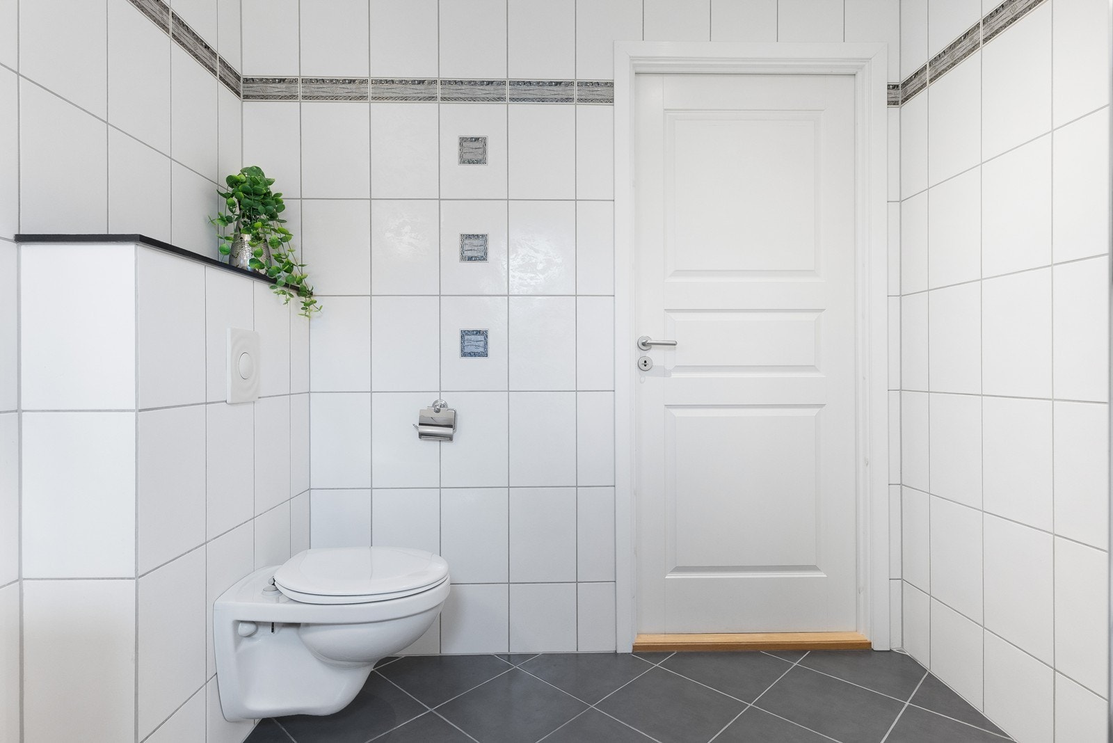 Badet har vegghengt toalett og gjennomført, tidløs utførelse med lyse fliser og mørke gulv for en stilren kontrast. Galleribilde