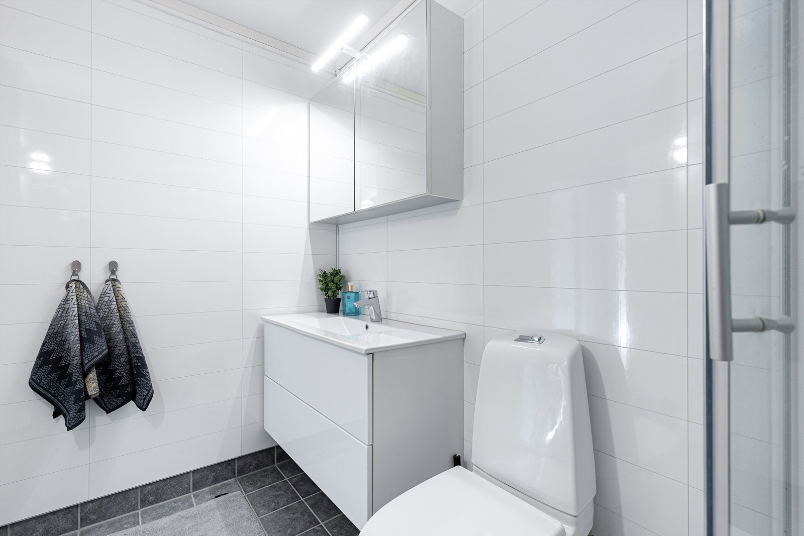 Badet er utstyrt med gulvmontert toalett, dusjkabinett og vegghengt servantinnredning, samt praktisk speilskap med overlys. Galleribilde