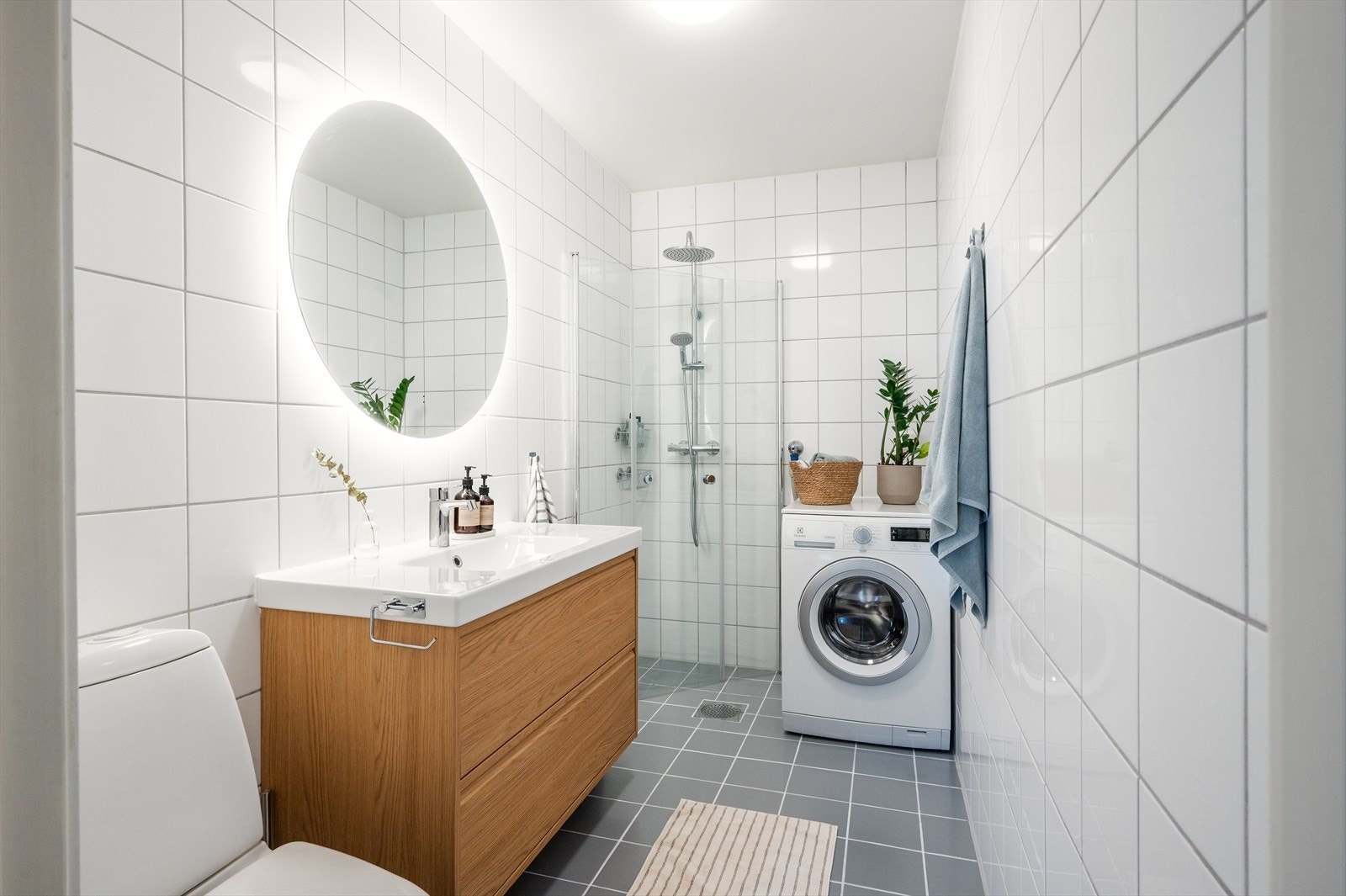 Pent og delikat flislagt bad med moderne uttrykk. Badet er innredet med toalett, servant med tilhørende innredning, speil med belysning samt dusjhjørne med svingbare glassdører. Galleribilde
