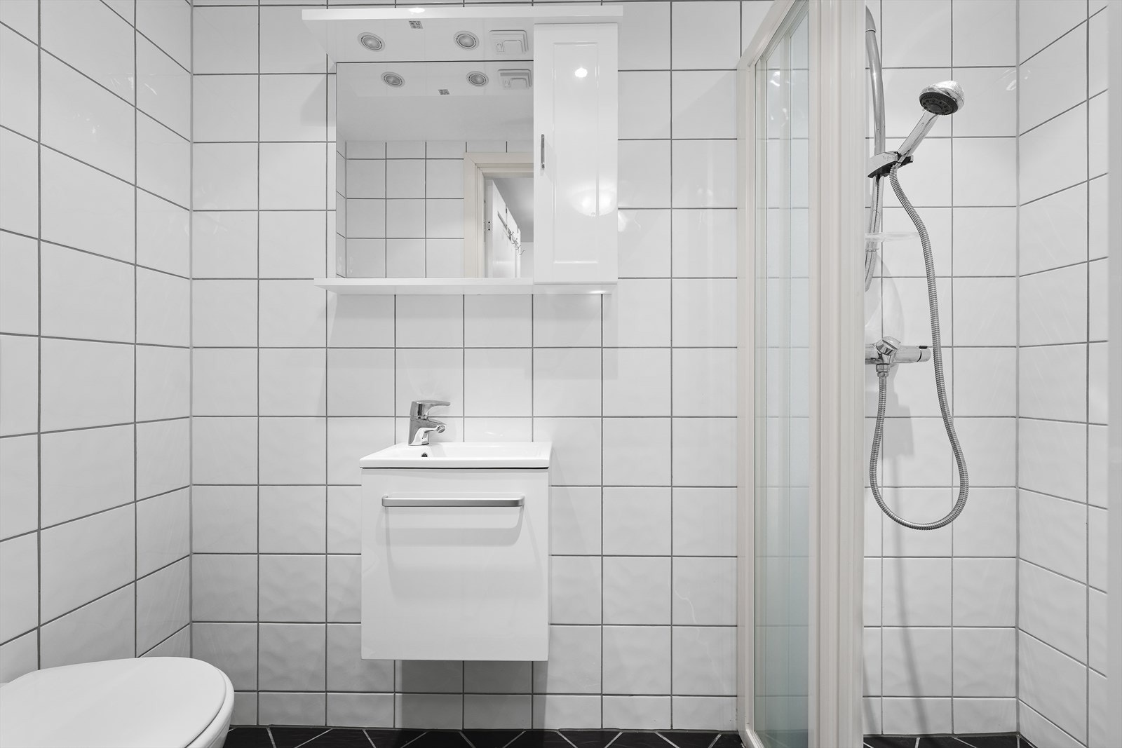 Flislagt baderom med gulvvarme. Badet er utstyrt med servant over innredning, speil med sideskap, dusjnisje og vegghengt toalett Galleribilde
