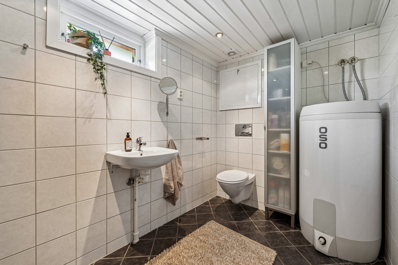 Baderom med vegghengt wc, servant og dusj i hjørne med innadslående dører og opplegg for vaskemaskin. Varmtvannstanken er på ca. 200 liter. fra 2023 plassert på kjellerbad. Galleribilde