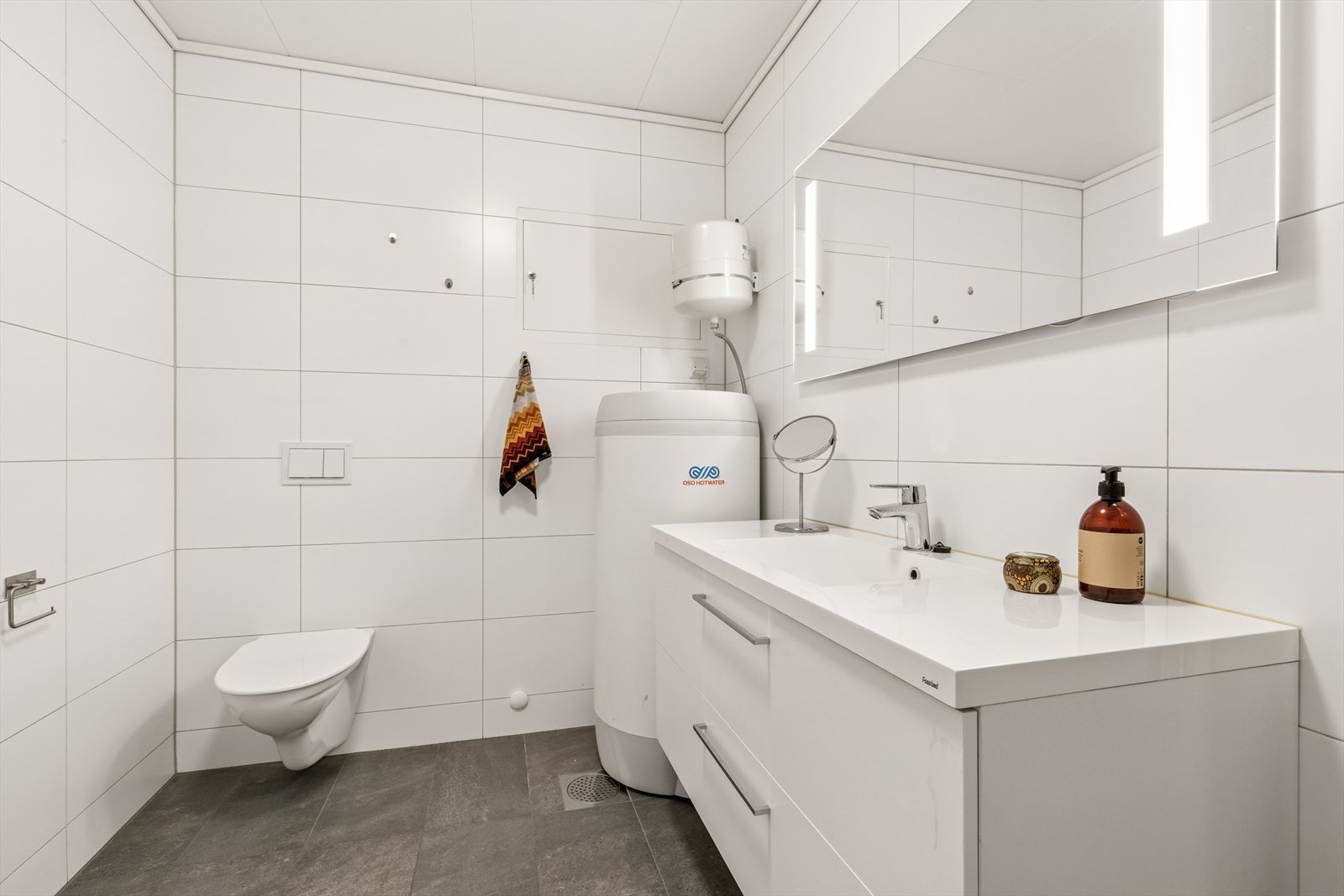 Badet er helfliset og fremstår moderne, utstyrt med servant, vegghengt toalett, dusj og varmtvannsbereder. Galleribilde