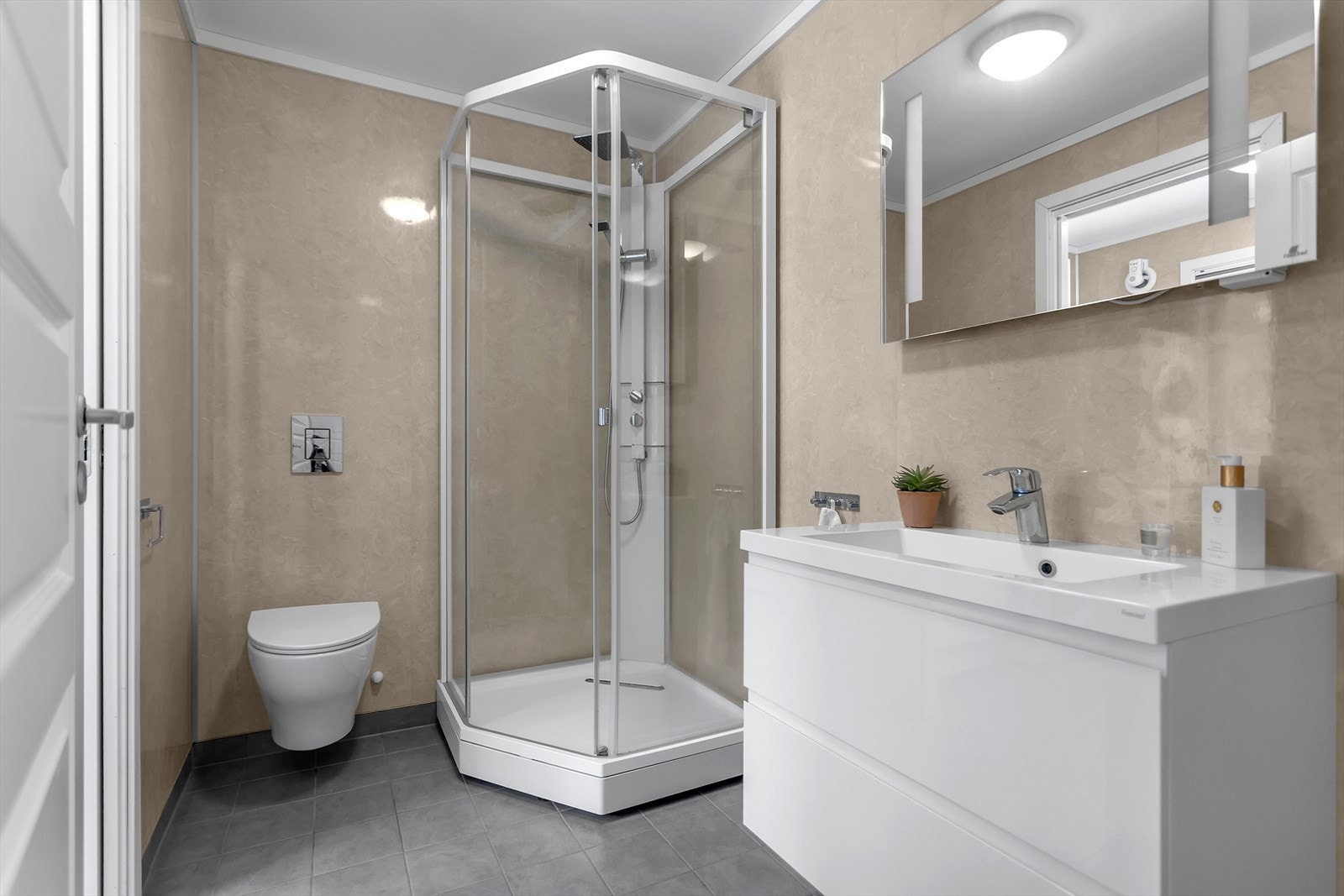 Det andre badet i hovedetasjen er designet for å kombinere komfort og praktiske løsninger. Rommet er innredet med et moderne dusjkabinett, et elegant speil med belysning, servant med romslige underskuffer og et stilrent vegghengt toalett. Galleribilde