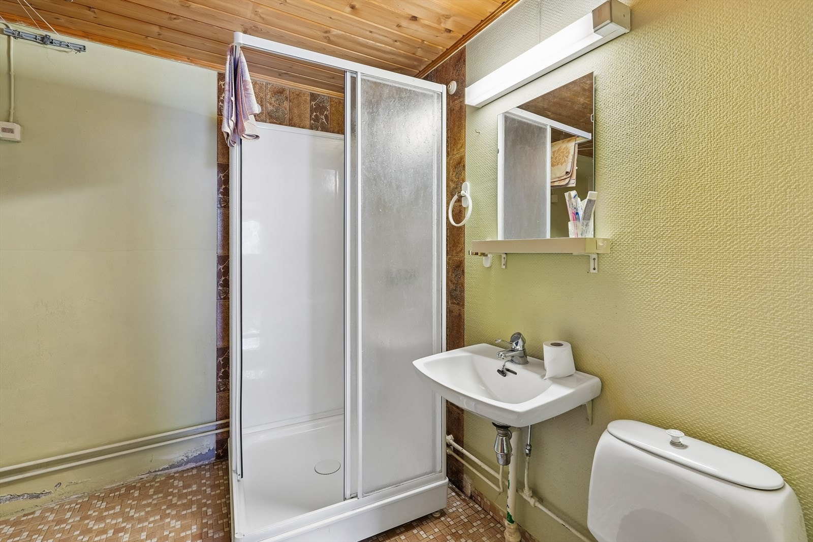 Badet i kjeller er også fra byggeår og er utstyrt med dusjkabinett, servant og toalett. Veggene har pussede og malte overflater kombinert med plater, mens gulvet er belagt med belegg. Galleribilde