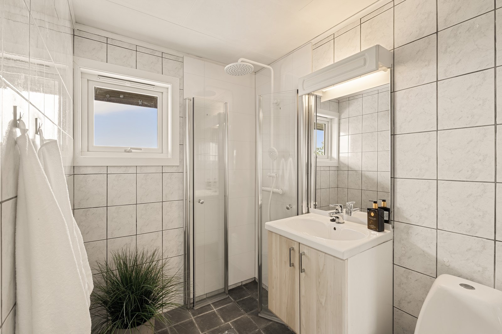 Badet er praktisk plassert i tilknytning til soverommene og er innredet med servant med møblement og speil, belysning, toalett, dusjgarnityr og dusjhjørne. Galleribilde