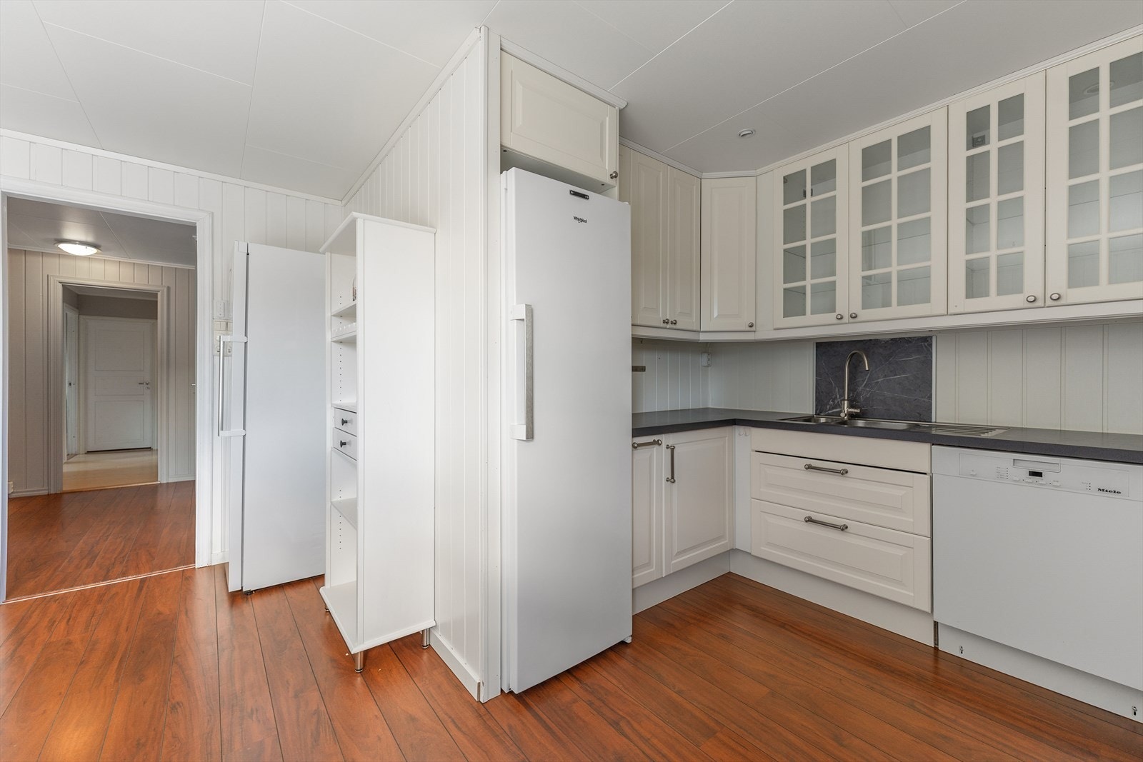 Kjøkken med lyse profilerte fronter og benkeplate av laminat. Kjøkkenet har innredning med profilerte fronter. Det er kjøleskap, oppvaskmaskin og komfyr. Det er kjøkkenventilator med avtrekk ut. Galleribilde
