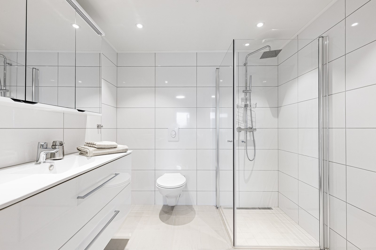 Badet er pent med flis på gulv og vegger. Her er det dusjhjørne med glassdører, vegghengt toalett og innredning med heldekkende hvit vask, underskuffer med slette fronter og overskap med speildører. Håndkletørker, varmekabler og balansert ventilasjon. Galleribilde