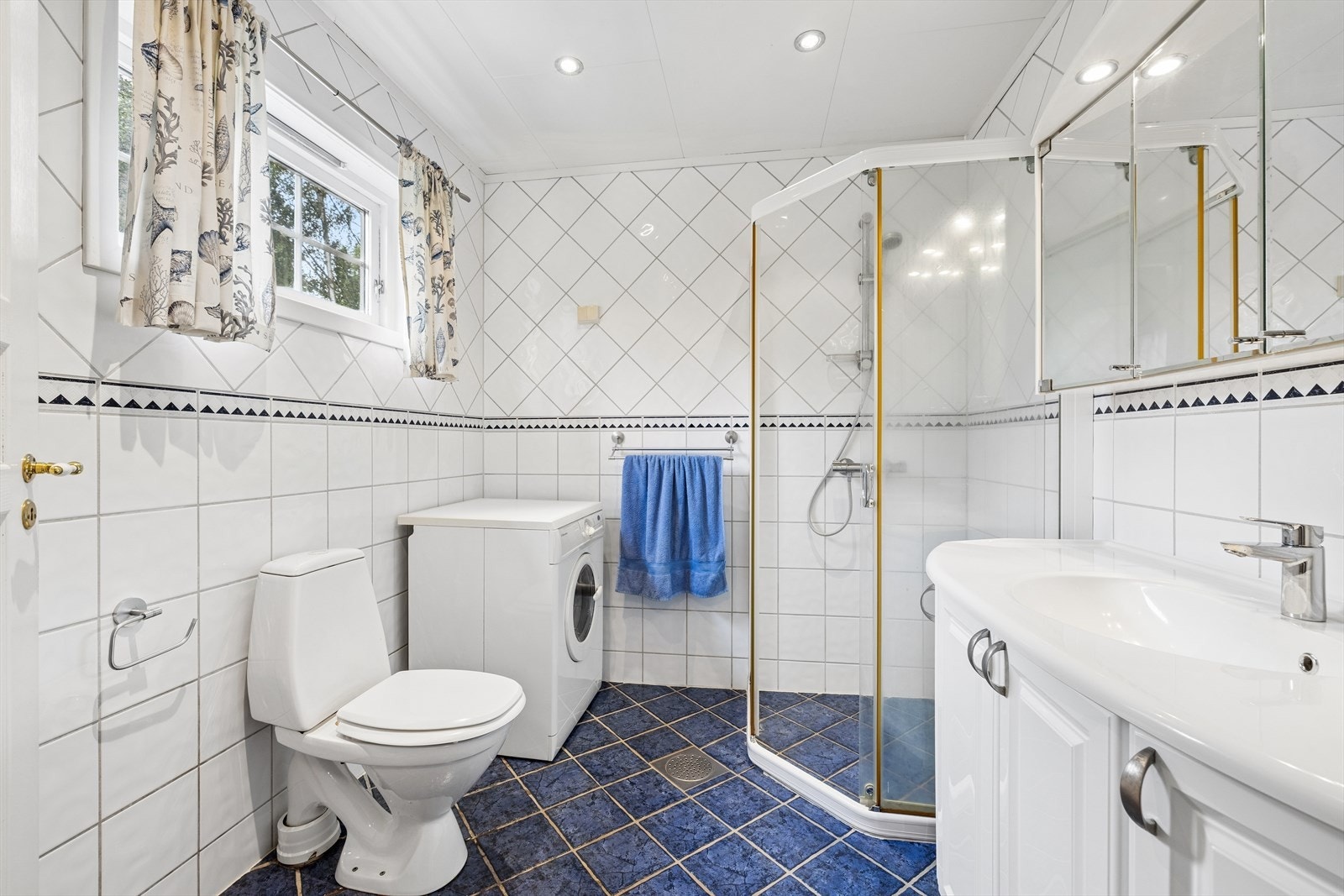 Badet er utstyrt med toalett, dusjhjørne med glassvegger, baderomsinnredning med heldekkende servant og speilskap, samt opplegg for vaskemaskin. Galleribilde