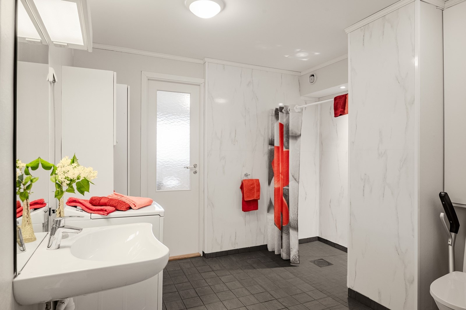 Badet har flislagt gulv og baderomsplater og malt tapet på veggene. Det er utstyrt med gulvmontert, handikaptilpasset toalett og romslig dusj med veggmontert dusjbatrteri. Galleribilde