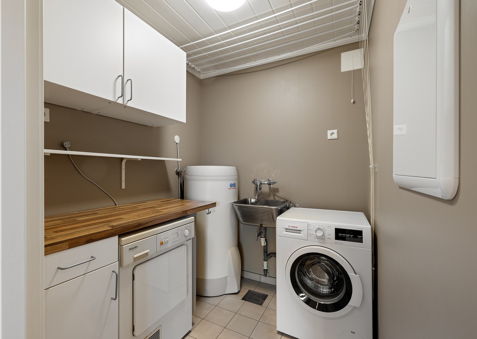 Praktisk og funksjonelt vaskerom med flislagt gulv. Innredningen består av under- og overskap og et praktisk tørkestativ er montert i taket. Det er opplegg for både vaskemaskin og tørketrommel. Galleribilde