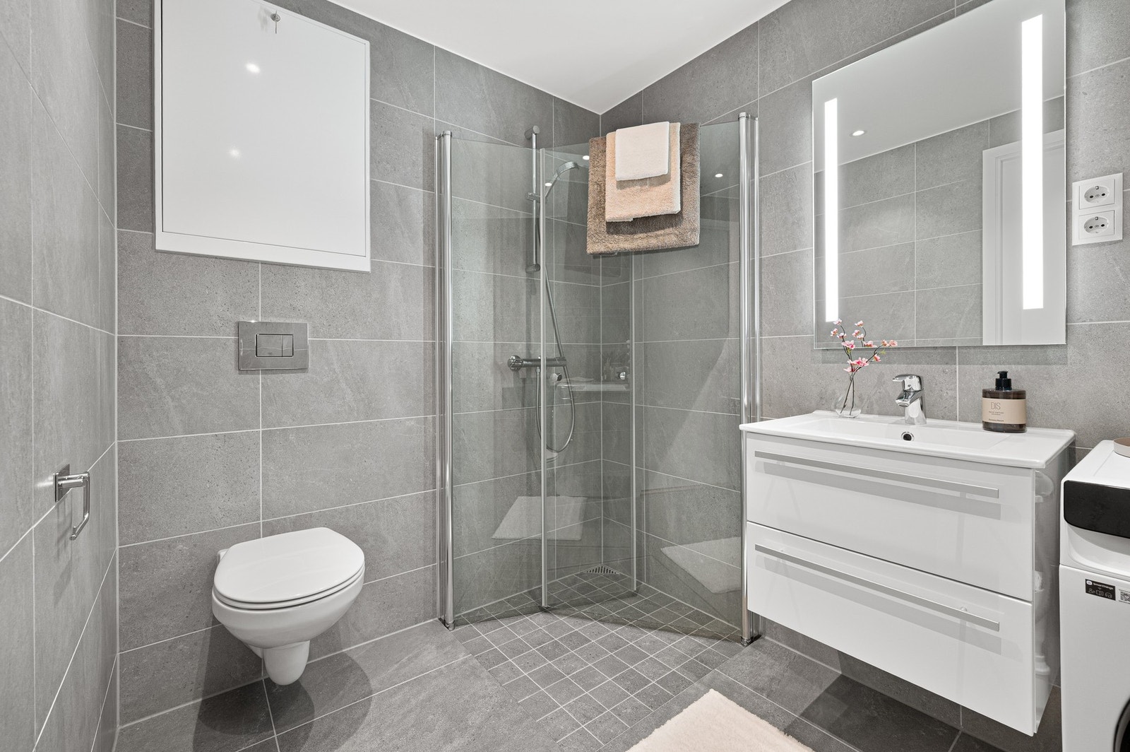 Badet er innredet med innfellbare dusjdører i glass, veggmontert WC, opplegg for vaskemaskin, servant på underskap, og speil. Galleribilde
