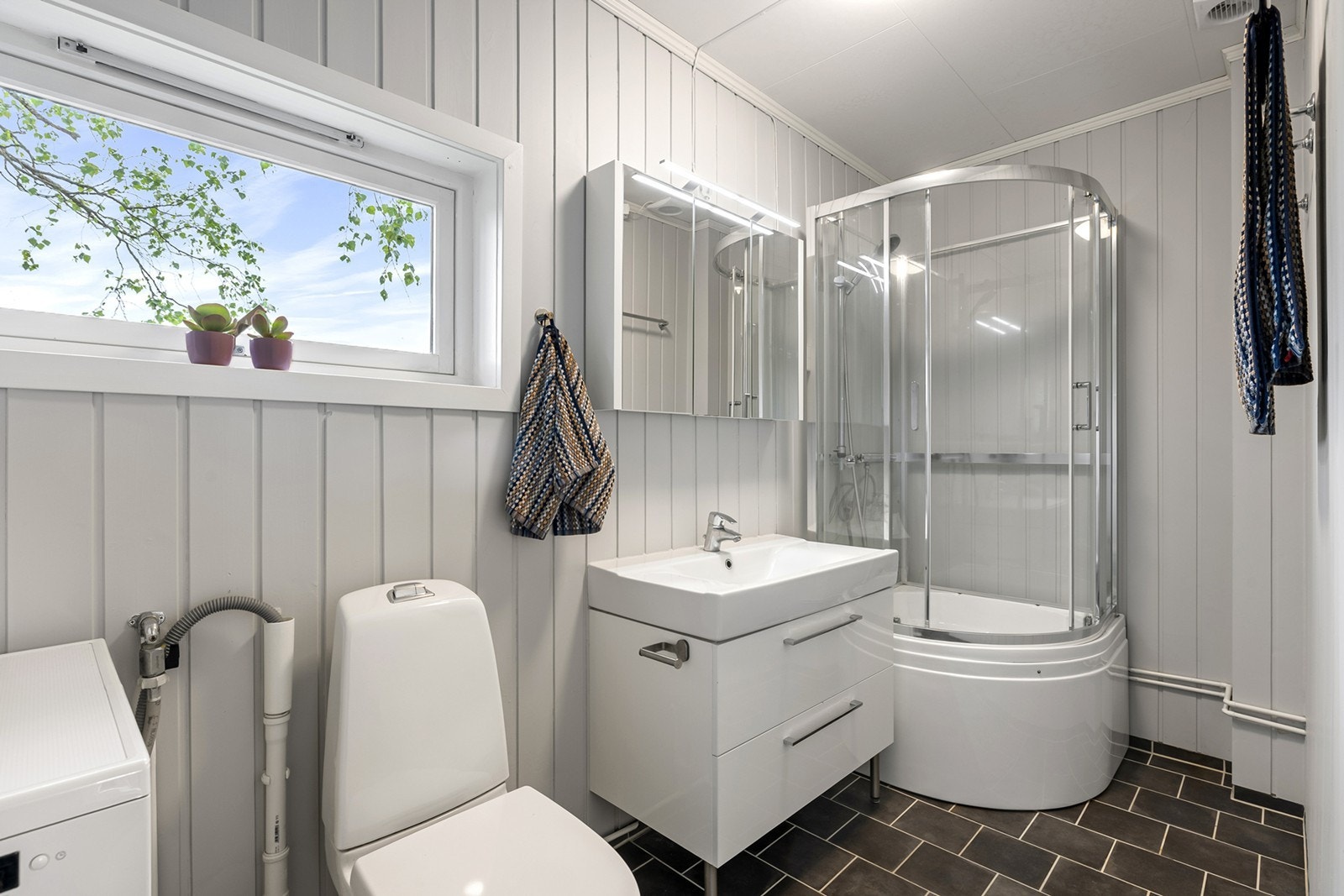 Badet har en lys og tidløs utforming med panel på veggene og fliser på gulvet, og er innredet med moderne servantskap, dusjkabinett og det er opplegg for vaskemaskin. Galleribilde