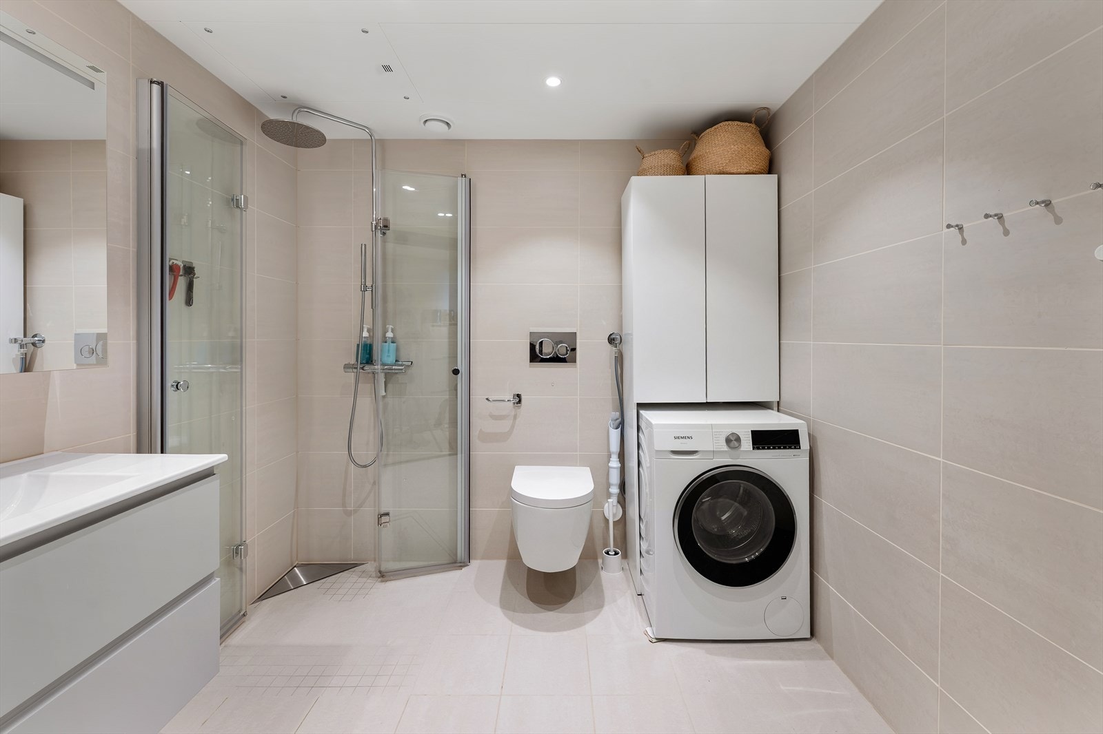 Badet fremstår som moderne og delikat, med lyse fliser på gulv med underliggende varme og vegger som gir rommet et tidløst og behagelig uttrykk. Badet er innredet med regnfallsdusj m/innfellbare glassdører, vegghengt toalett og opplegg for vaskemaskin Galleribilde