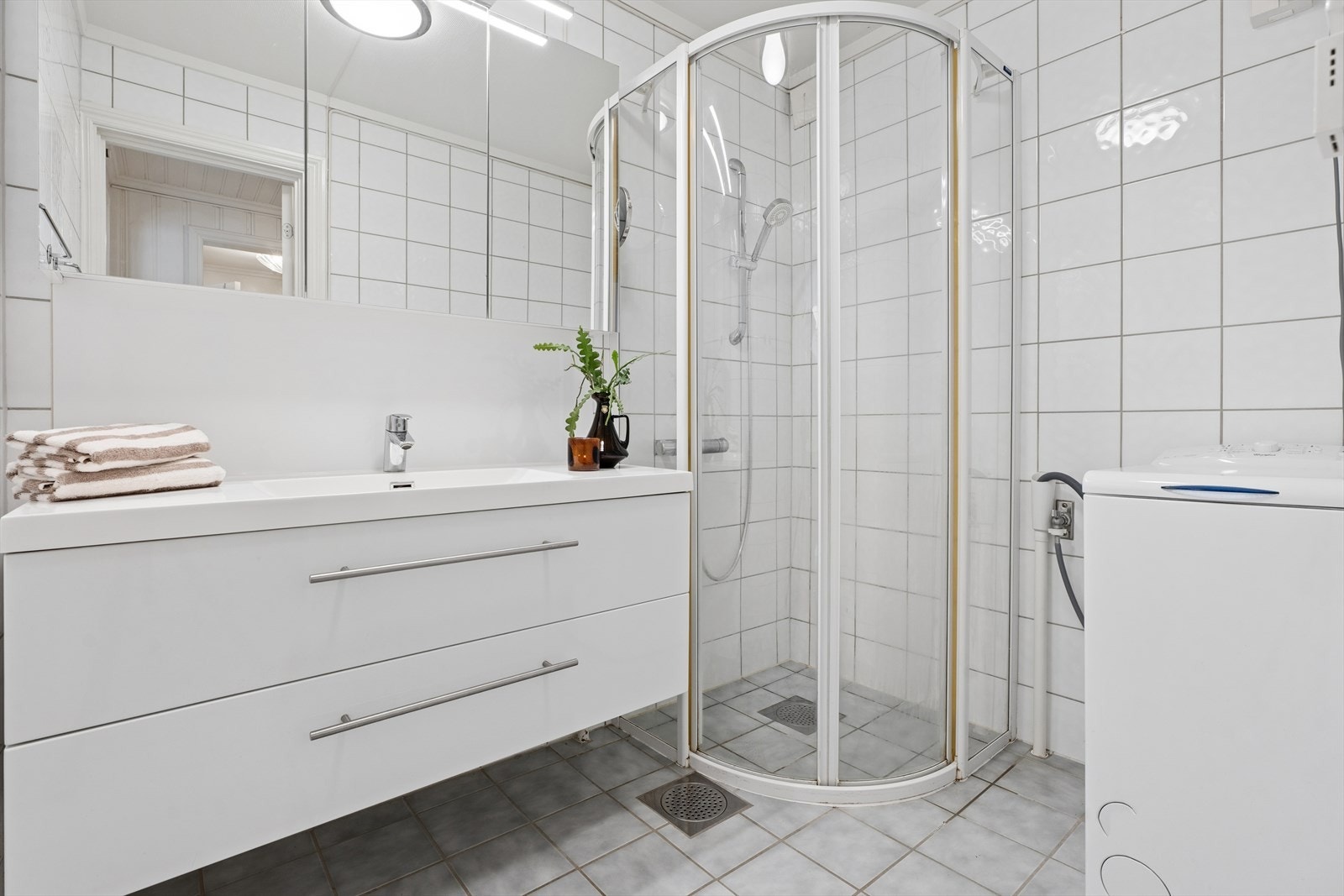 Badet er utstyrt med dusjhjørne, servant med underskap og opplegg for vaskemaskin. Det er varme i det flislagte gulvet. Galleribilde