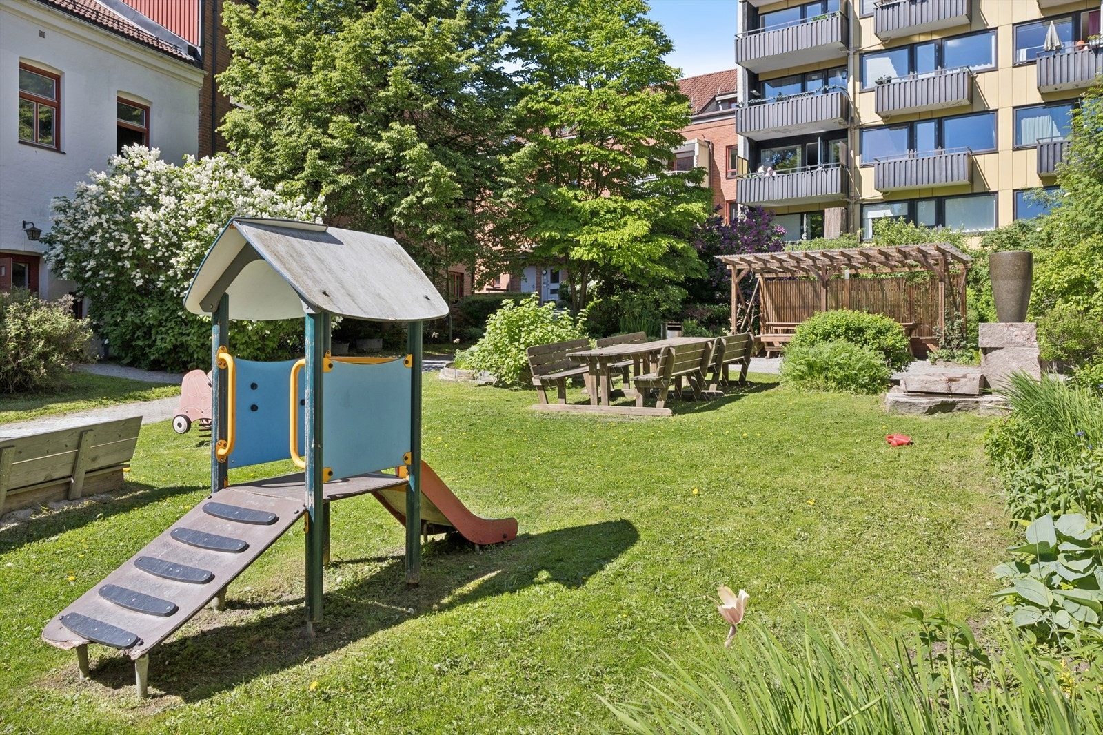 En grønn oase av en bakgård. Det er frodig beplantet med blomstrende busker og store trær, der en dekorativ steinskulptur med sildrende vann setter en rolig og nesten parkaktig stemning. Galleribilde