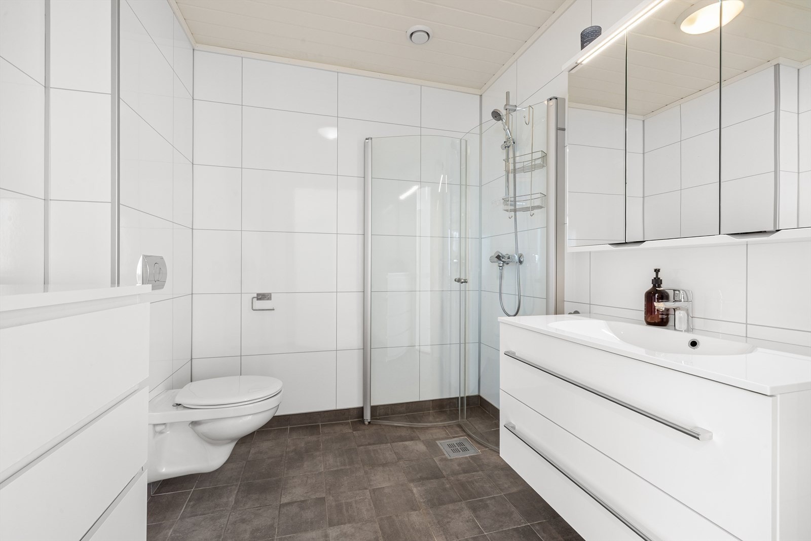 Badet i 2. etasje har samme stilnivå som badet i underetasjen, med flislagt gulv og Fibo veggpanel på vegger. Innredningen består av servant med skapløsning, speil, toalett og dusjhjørne med glassdører. Galleribilde