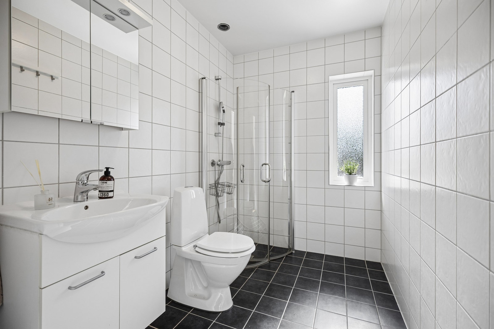 Flislagt bad med hvite vegger og grått gulv som gir et lyst og tidløst uttrykk. Badet har servantskap, speilskap med oppbevaring, speil med lys, WC og dusjhjørne med glassdører som kan legges inn. Gulvvarme gir god komfort året rundt. Galleribilde