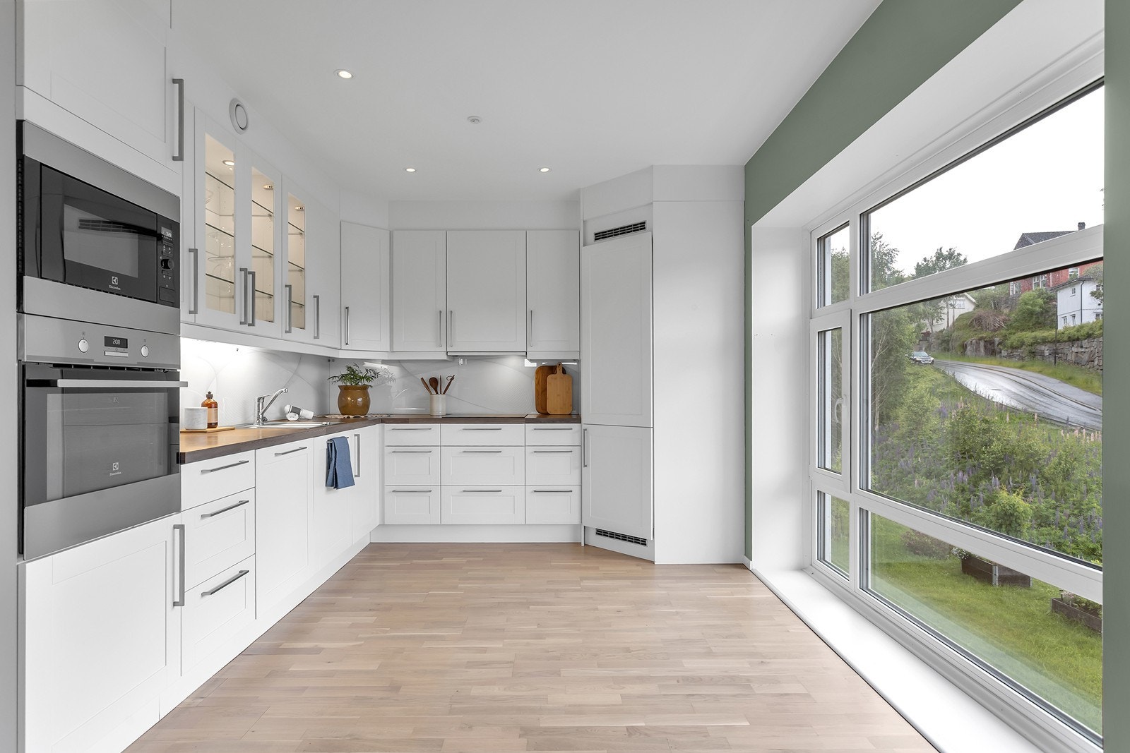 Lyst kjøkken med hvit innredning, glatte fronter og god skap- og benkeplass. Den store vindusflaten gir rikelig med naturlig lys og et hyggelig utsyn. Galleribilde