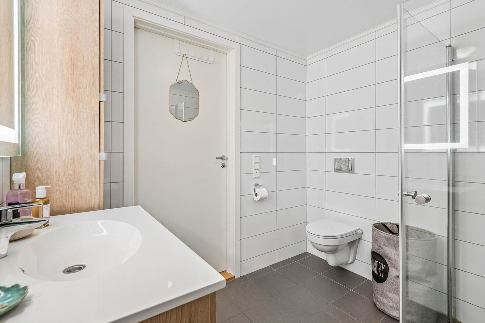 Flislagt baderom med behagelig gulvvarme. Badet er utstyrt med vegghengt toalett, dusjvegger av glass, servant med underskap, speil med lys, og høyskap. Galleribilde