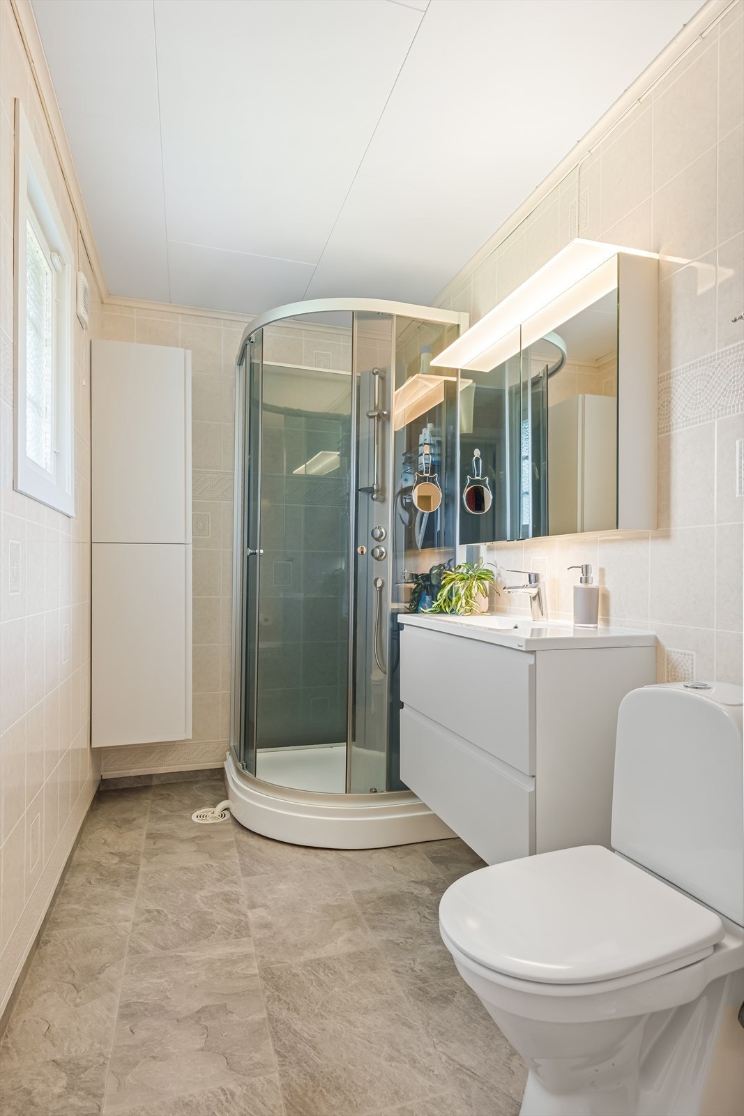 Lyst bad med dusjkabinett, servant, speil og gulvstående toalett. Badet er utstyrt med elektriske varmekabler, baderomsplater på vegg og belegg på gulvet. Galleribilde