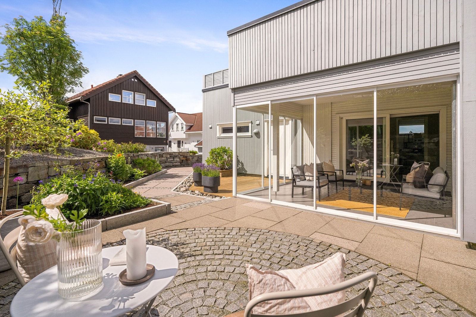 uteområdet pent opparbeidet med steinlagte partier, tredekke, blomstrende bed, natursteinsmurer og grønne innslag. Galleribilde
