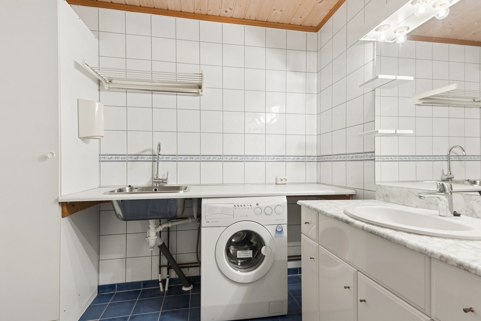Rommet er et kombinert bad og vaskerom, med opplegg for vaskemaskin og praktisk utslagsvask, i tillegg til innredning med nedfelt servant, toalett og dusj. Galleribilde
