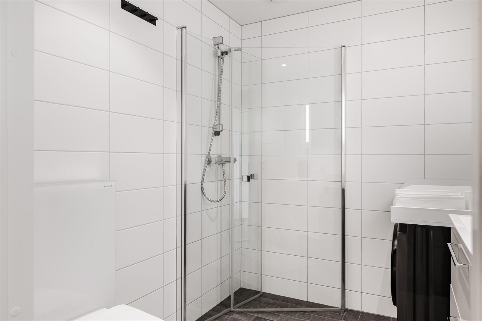 Baderommet er også utstyrt med dusjhjørne med innfellbare glassdører og veggmontert toalett. I hjørnet er det plass og opplegg for vaskemaskin, samt tørketrommel over om ønskelig. Galleribilde