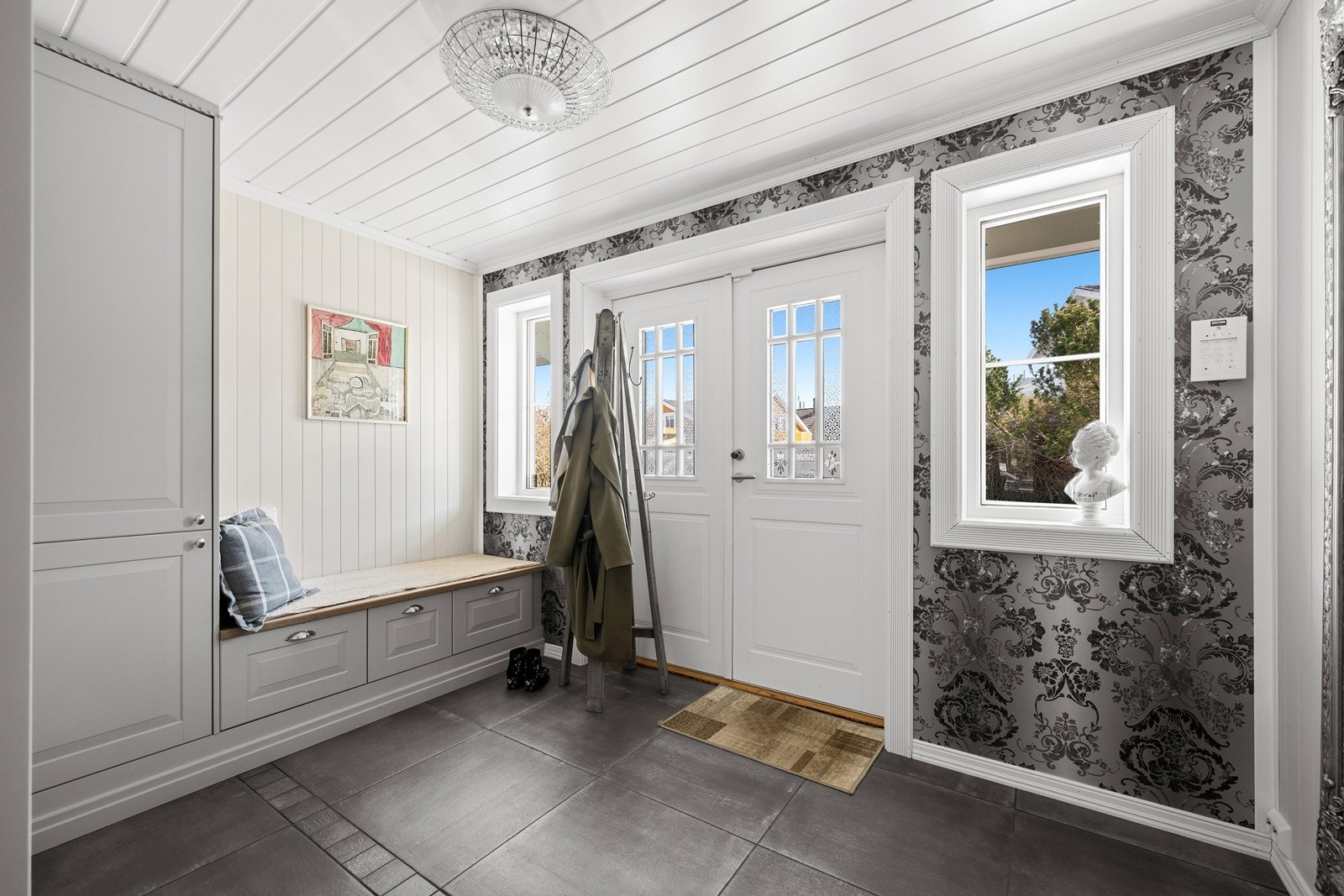 Romslig entré med grå fliser på gulvet, doble dører og rikelig med naturlig lys. Veggene er utført i en kombinasjon av tapet og malt panel. Galleribilde