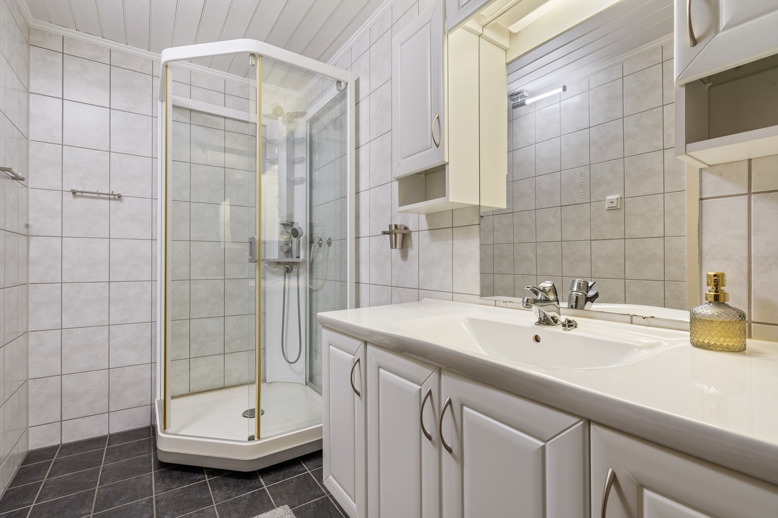 Badet i underetasjen har fliser på vegg og gulv. Panel i tak. Rommet inneholder et tett dusjkabinett, seksjon med servant og toalett på sokkel. Galleribilde