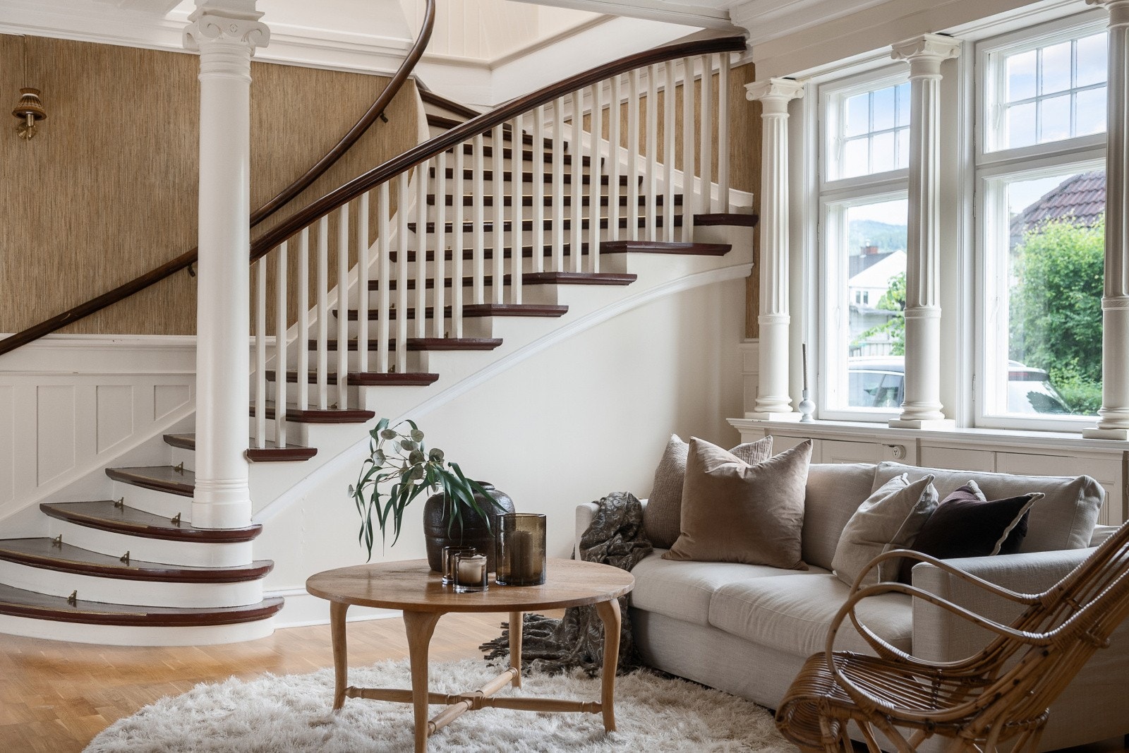 En svungen trapp med mørklakkert rekkverk og en søyle i tre stiger fra hallgulvet opp mot 2. etasje _ en konstruksjon som bærer preg av håndverk fra en annen tid. Galleribilde