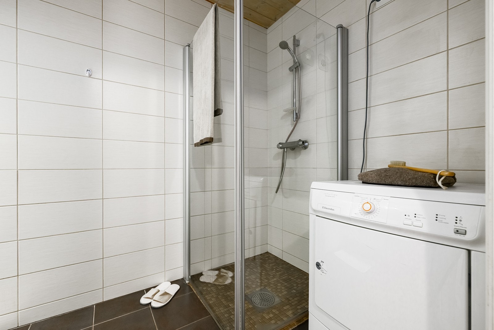 Badet fremstår som funksjonelt og velholdt med flislagte overflater, dusjhjørne, vegghengt toalett, baderomsinnredning og opplegg for vaskemaskin. Galleribilde