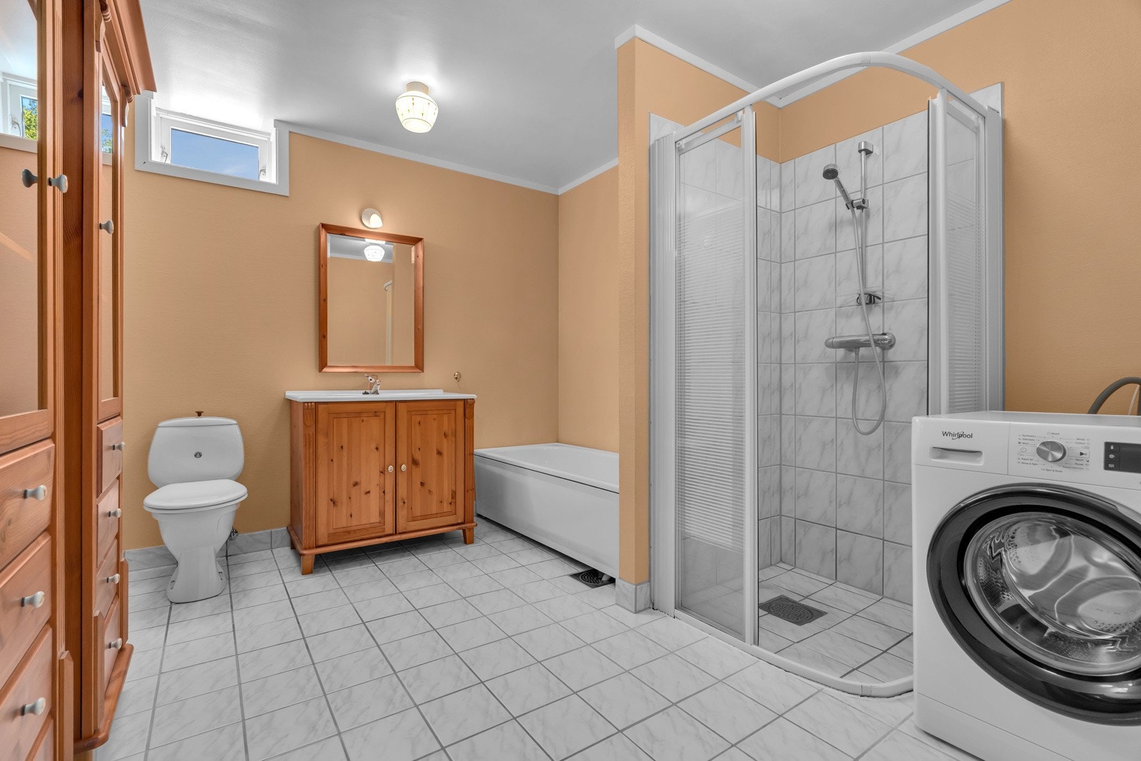 Badet er lyst med fliser med varmekabler i gulv, malte vegger, badekar, dusjhjørne og opplegg til vaskemaskin. Galleribilde