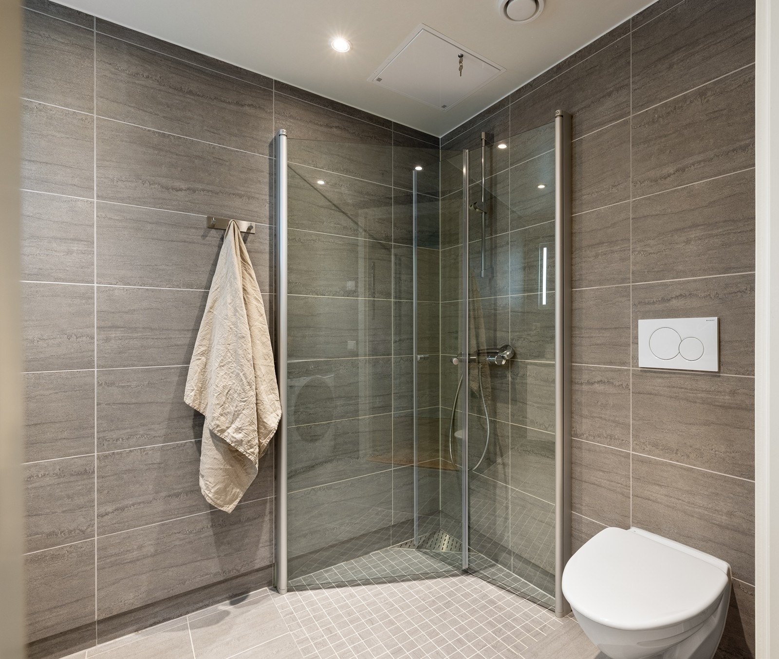 Badet er stilrent innredet med en heldekkende skuffeservant, speil med integrert belysning, et dusjhjørne med innfellbare glassvegger, samt et moderne vegghengt toalett med skjult sisterne. Galleribilde