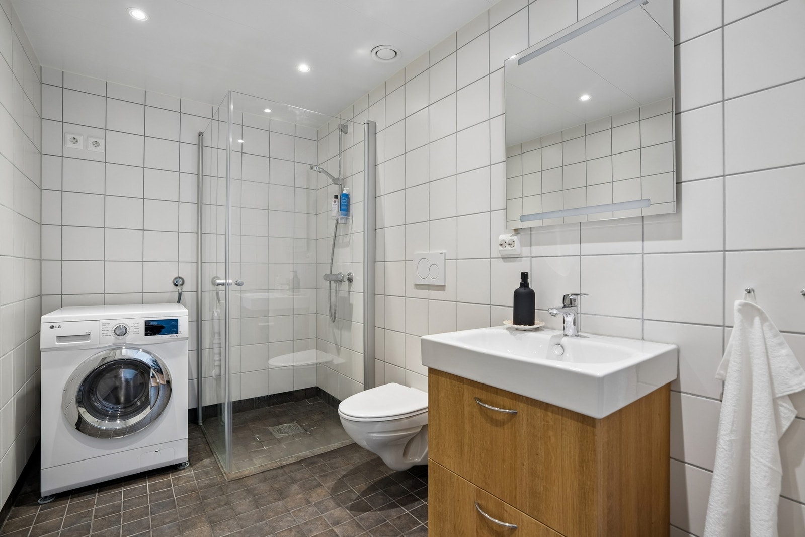 Tidløst og pent flislagt bad med varme i gulvet. Badet er innredet med vegghengt toalett, dusjhjørne, servant på benkeskap samt speil med integrert belysning. Galleribilde